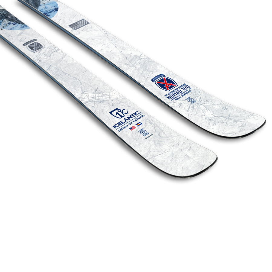 Two white Icelantic Skis, the 10th Mtn Nomad 100 model, featuring blue and red logos—including the Icelantic and National Ski Patrol emblems—are displayed flat near the tips against a white background. This special edition ski celebrates alpine heritage by commemorating the 10th Mountain ski legacy while delivering performance inspired by the original NOMAD 100.
