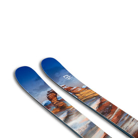 A close-up of two blue Icelantic Skis Arches NP 100 freeride skis, showcasing a painted landscape of red rock formations and a cloudy sky, with a small white mountain logo on one ski.
