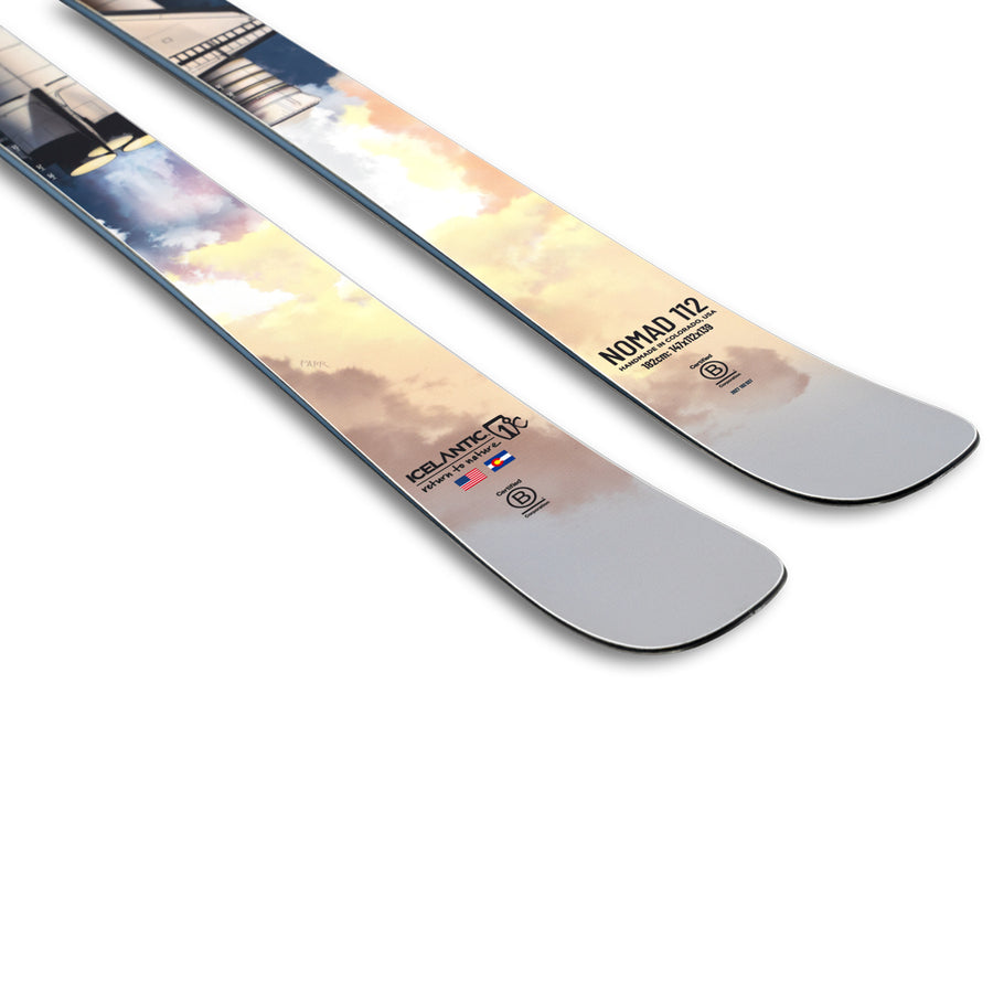 Close-up of two light-colored Icelantic Skis with a pastel sky design, showing the tips. The ski on the right is labeled Nomad 112 and features several small logos and symbols near the tip.