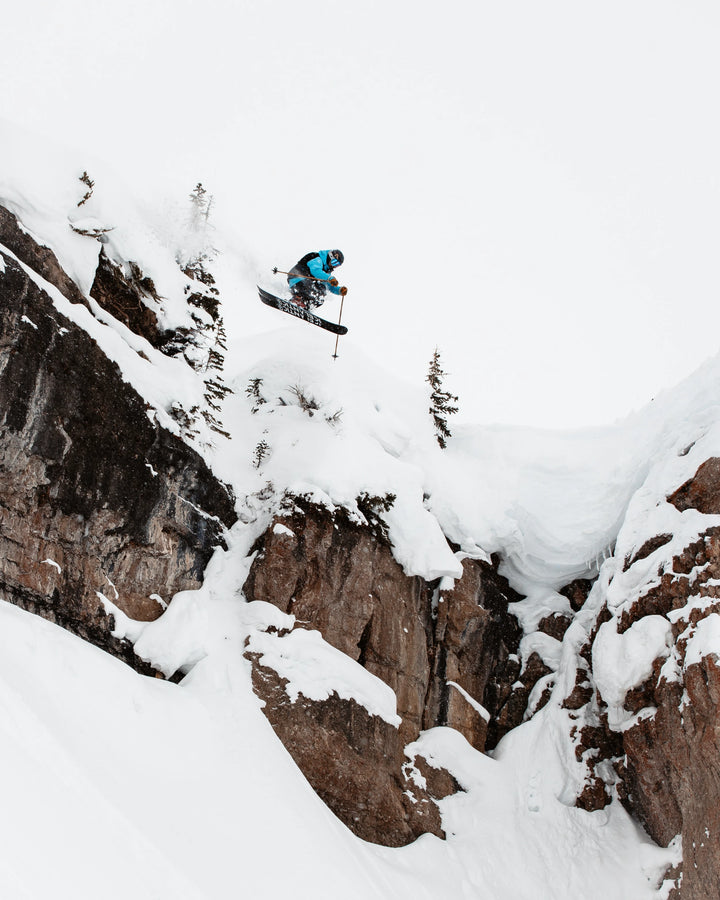 Clad in blue gear, a skier launches off a snowy cliff on Icelantic Skis’ Nomad 106, soaring high above the ground with rocky ledges and snow-dusted trees behind them.