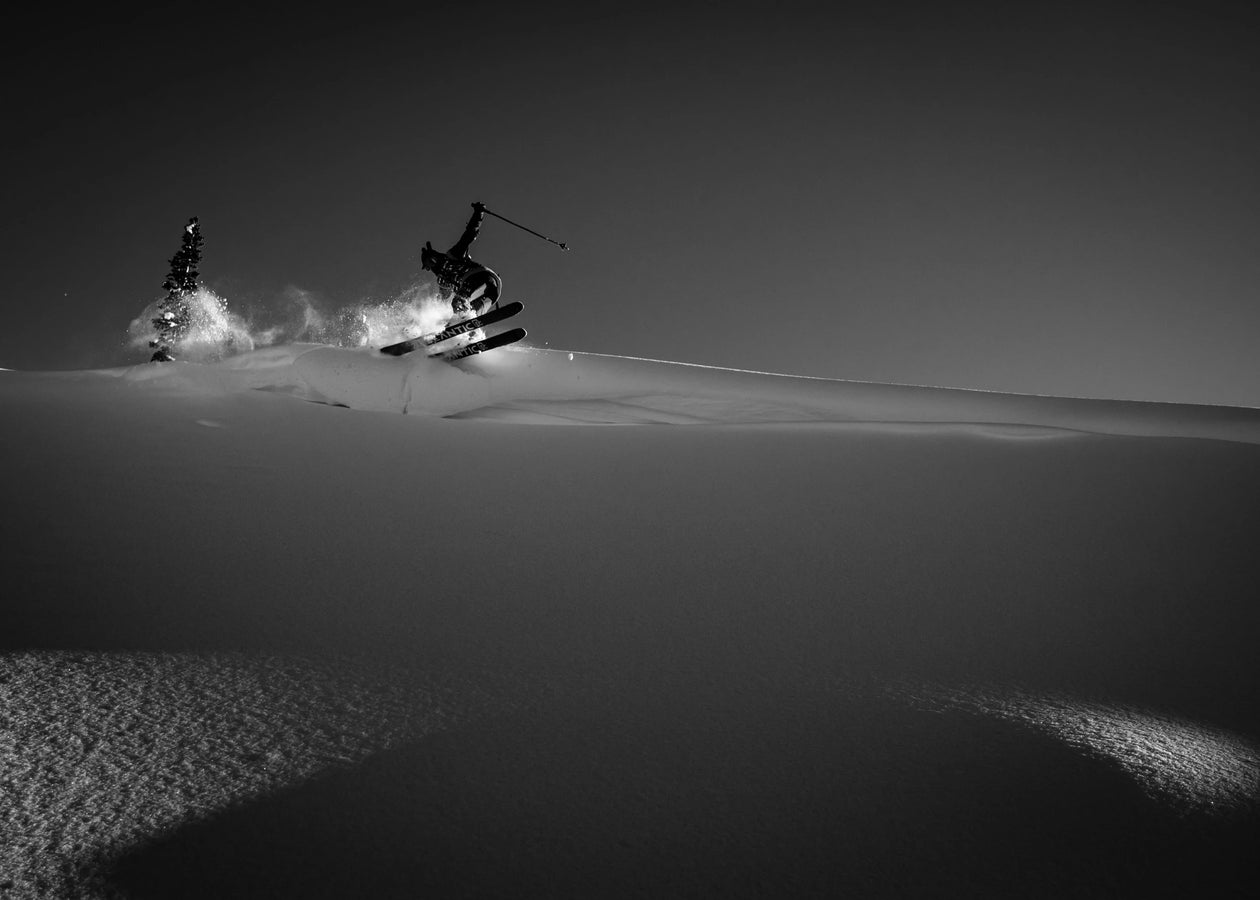 An athlete soars through the air on a snowy slope, launching off a jump with snow spraying behind, riding the Icelantic Skis Nomad 106. The striking black and white scene emphasizes the bold contrast between the skier and the winter landscape.