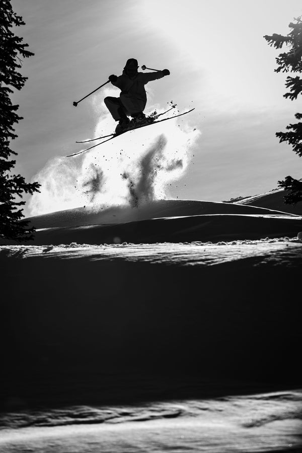 A skier launches off a snowy slope on Icelantic Skis’ Nomad 100, their silhouette outlined against the bright sky as snow sprays behind them. Pine trees border the black and white scene.