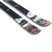 Two Icelantic Skis Maiden 108 skis with colorful designs showcasing a person’s face wearing ski goggles and a helmet, snowy mountains, a building, and a starry sky in the background.