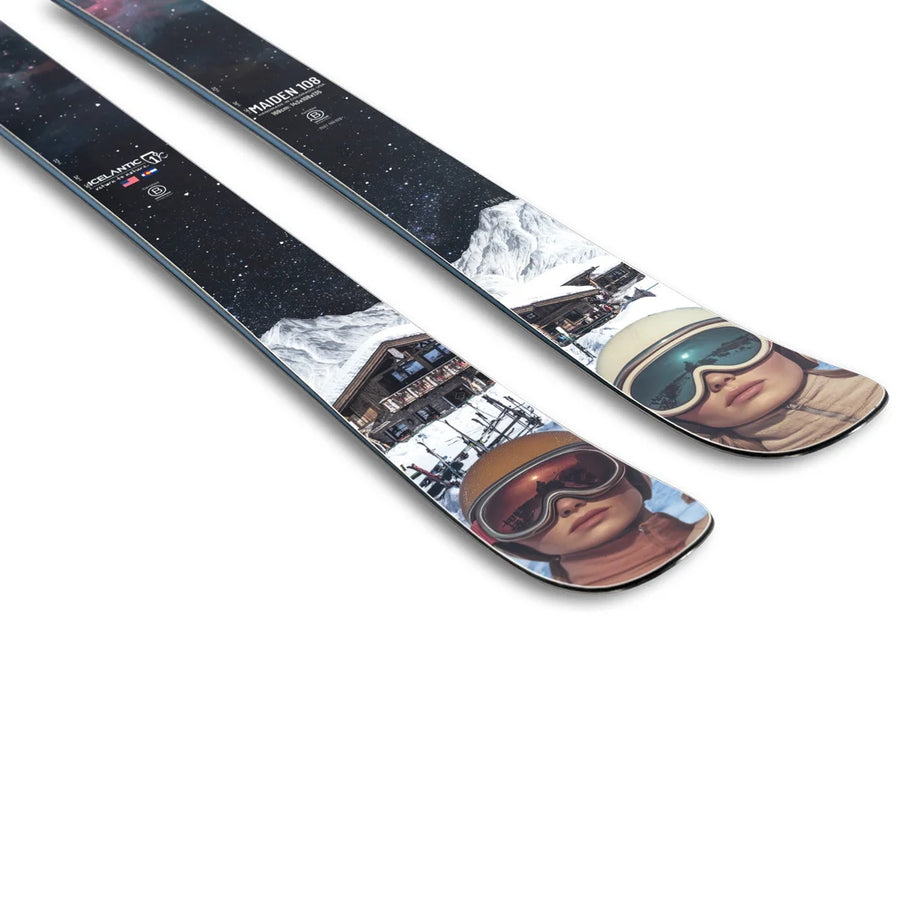 Two Icelantic Skis Maiden 108 skis with colorful designs showcasing a person’s face wearing ski goggles and a helmet, snowy mountains, a building, and a starry sky in the background.