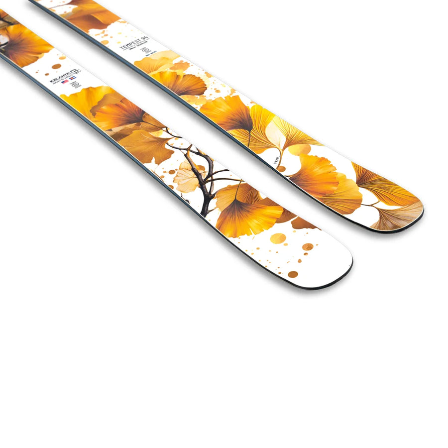 A close-up of two Icelantic Skis Tempest 94 white skis, showcasing a yellow and orange ginkgo leaf pattern with branches and abstract dots.