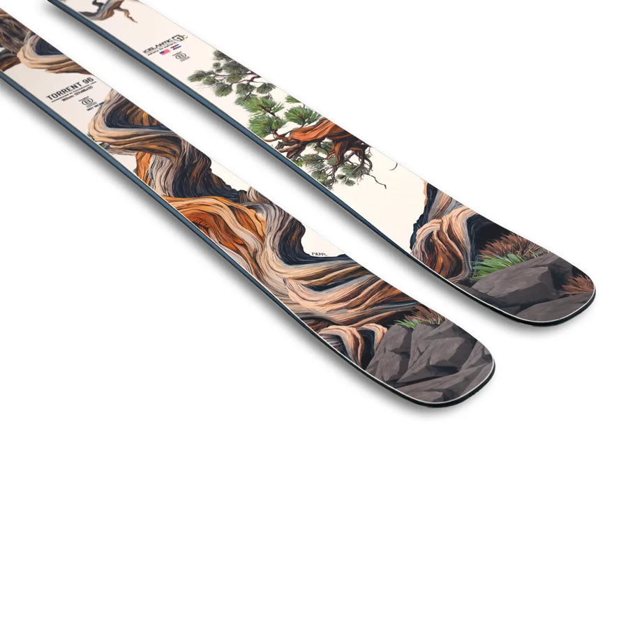 A close-up of two Icelantic Skis Torrent 96 skis showcases artistic designs of twisted trees, rocks, and greenery on a white background, with detailed artwork adorning the upper surfaces near the tips.