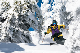 A skier glides swiftly through fresh snow between snow-covered trees on a sunny winter day, carving turns with the Maiden 102 from Icelantic Skis.