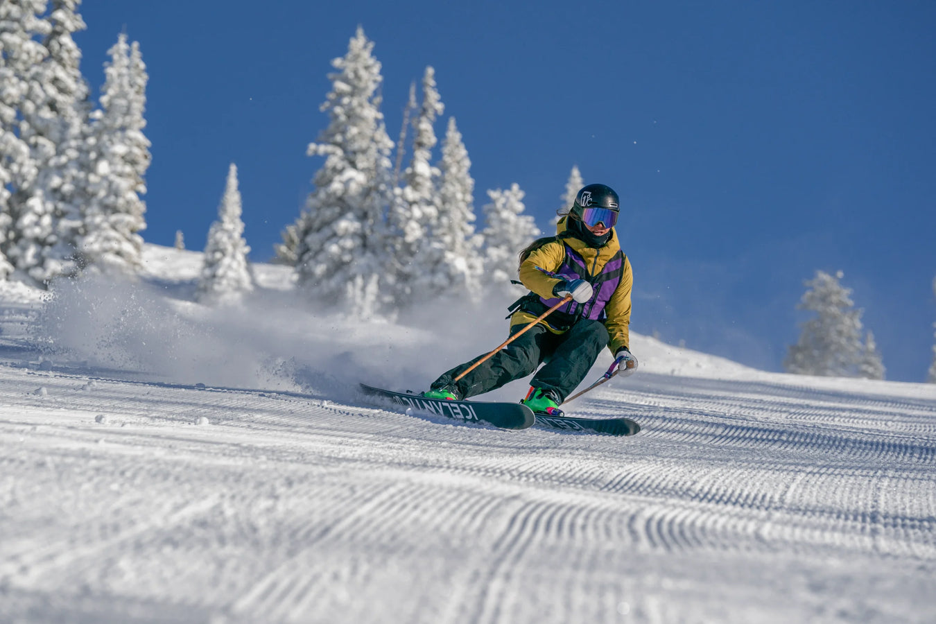 Riding the Icelantic Skis Maiden 94, a skier in a yellow jacket and green pants carves down a groomed snowy slope, framed by snow-covered trees and a clear blue sky.