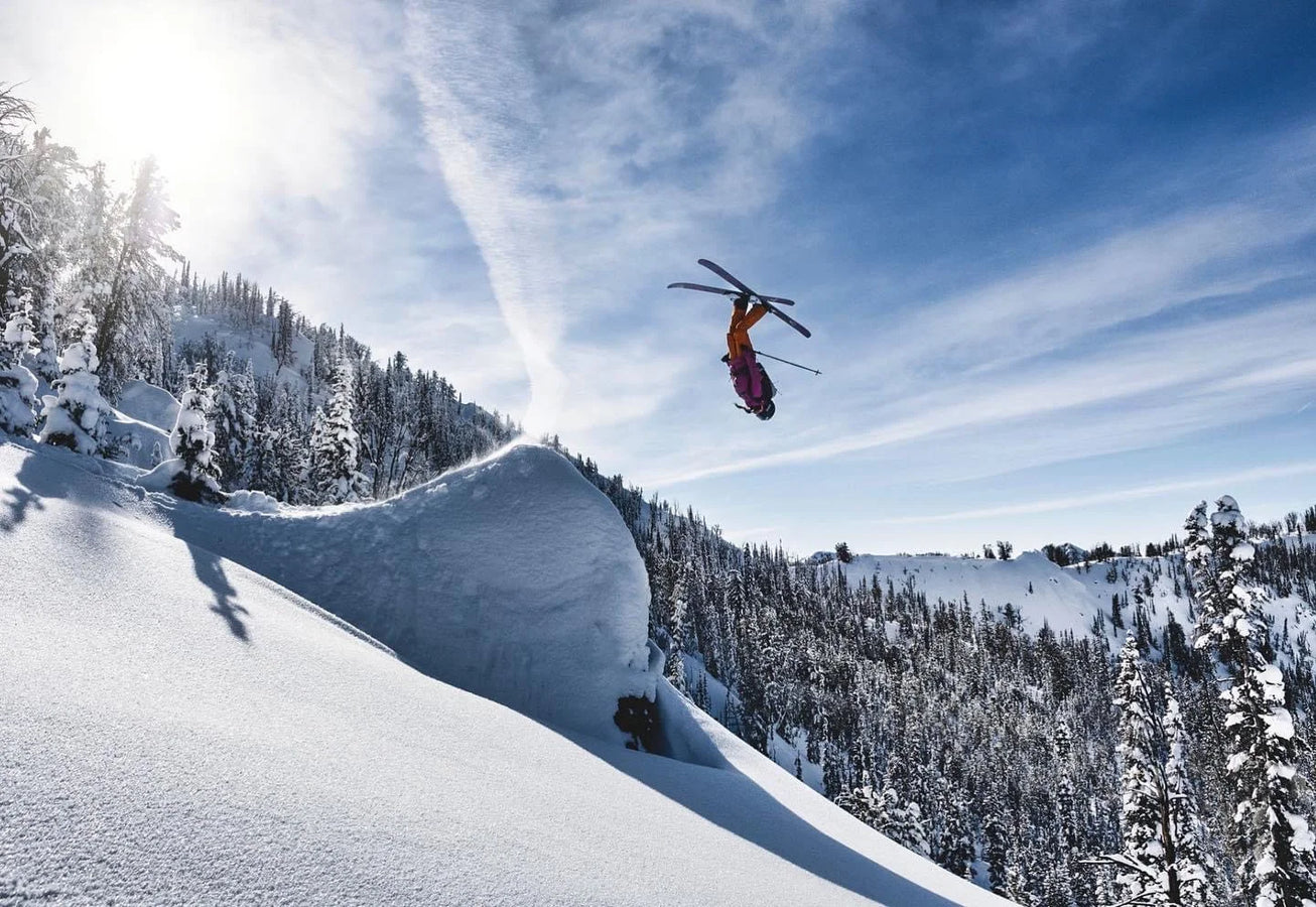 Soaring through the air on Icelantic Skis’ Maiden 102, a skier executes a backflip above a snowy slope framed by snow-laden trees and a vivid blue sky.