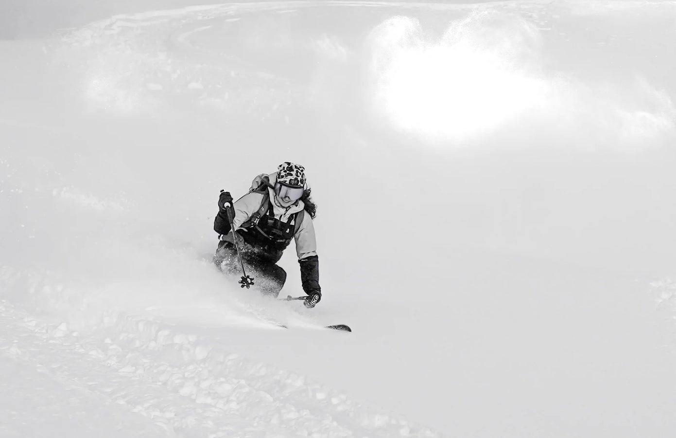 A skier in winter gear is carving through deep, fresh snow on a mountain slope with the Icelantic Skis Maiden 102, creating a spray of powder behind them under bright sunlight.