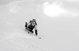 A skier in winter gear is carving through deep, fresh snow on a mountain slope with the Icelantic Skis Maiden 102, creating a spray of powder behind them under bright sunlight.