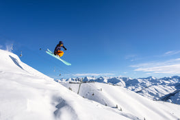 A skier on Icelantic Skis’ Maiden 94 launches off a snowy slope in orange pants under a clear blue sky, with mountain peaks and a ski lift visible in the background.