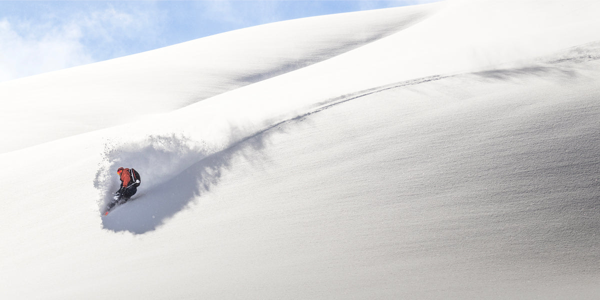 A person on a snowmobile speeds through deep, untouched snow on a sunny, white mountain slope, creating a spray of powder behind them.