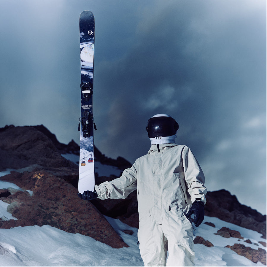 Clad in a white spacesuit and helmet, a figure stands on a snowy, rocky slope beneath dramatic, cloudy skies, holding an Icelantic Skis Nomad 106 upright—evoking the feeling of exploring an otherworldly and frigid landscape.