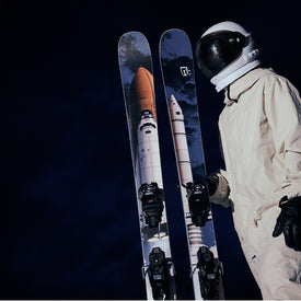 An individual in an astronaut suit holds Icelantic Skis’ Nomad 112 freeride skis, which are adorned with space shuttle graphics and set against a dark, night sky background.