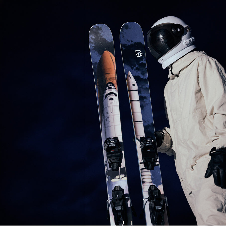 An individual in an astronaut suit holds Icelantic Skis’ Nomad 112 freeride skis, which are adorned with space shuttle graphics and set against a dark, night sky background.