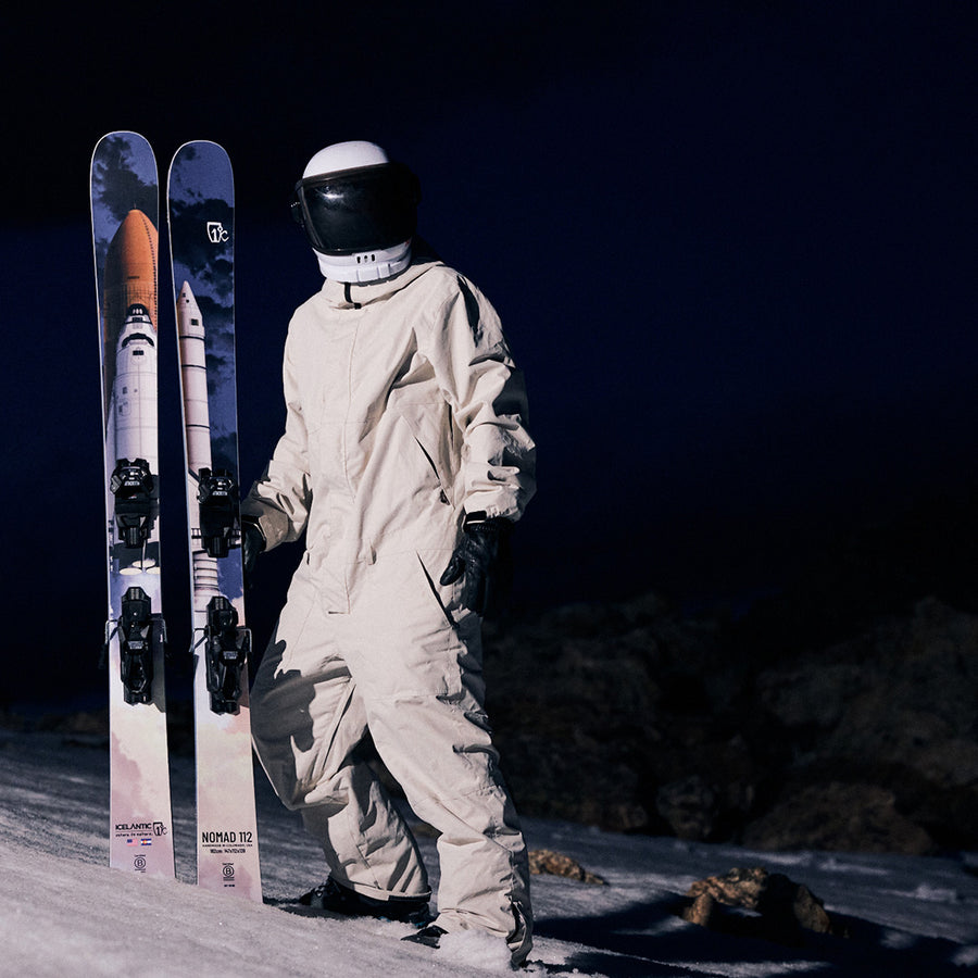 A person in a spacesuit stands on snow at night, holding a pair of Icelantic Skis Nomad 112 advanced freeride skis, which are decorated with images of a space shuttle against a dark sky.