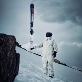 A person in a white spacesuit and black helmet stands on snowy ground, holding an Icelantic Skis Nomad 112 upright. Rocks and a cloudy sky are in the background, creating an adventurous scene that would appeal to advanced skiers.