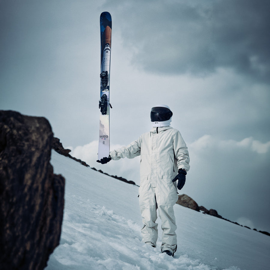 A person in a white spacesuit and black helmet stands on snowy ground, holding an Icelantic Skis Nomad 112 upright. Rocks and a cloudy sky are in the background, creating an adventurous scene that would appeal to advanced skiers.