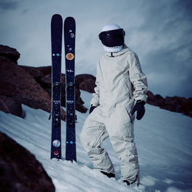 A person in a white spacesuit and helmet stands on snowy ground beside a pair of Icelantic Skis Nomad 94 all-mountain skis decorated with planet stickers, with rocky terrain in the background.