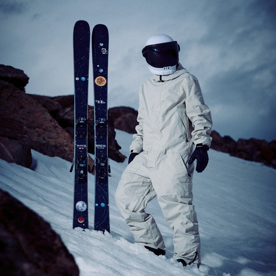 A person in a white spacesuit and helmet stands on snowy ground beside a pair of Icelantic Skis Nomad 94 all-mountain skis decorated with planet stickers, with rocky terrain in the background.