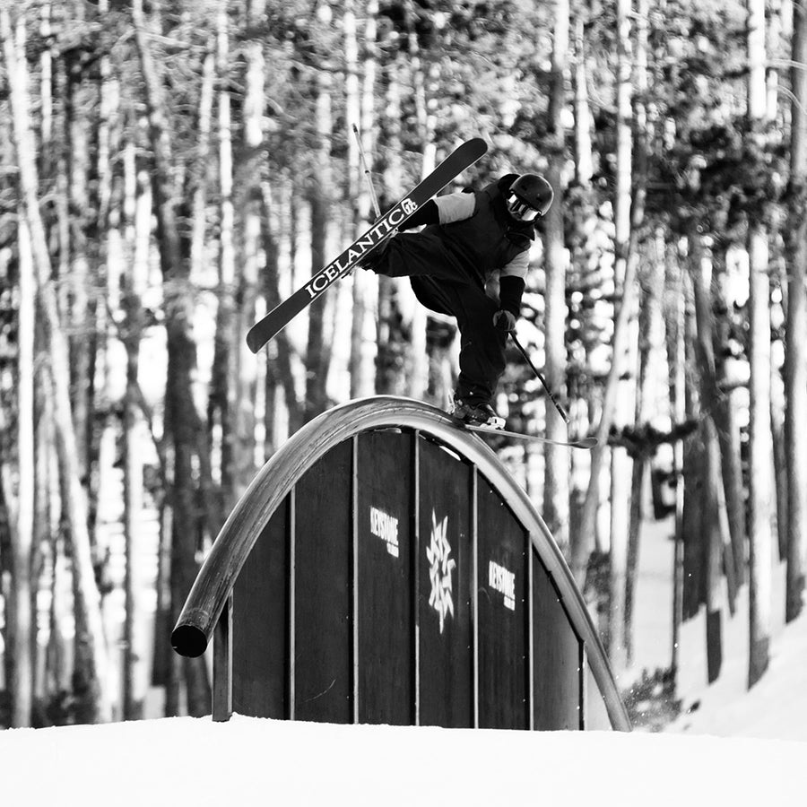 A skier riding the Icelantic Skis Nomad 94 Whiteout tackles a curved rail in a snowy forest, holding one ski and balancing mid-air. The black and white image captures the dynamic freestyle trick.