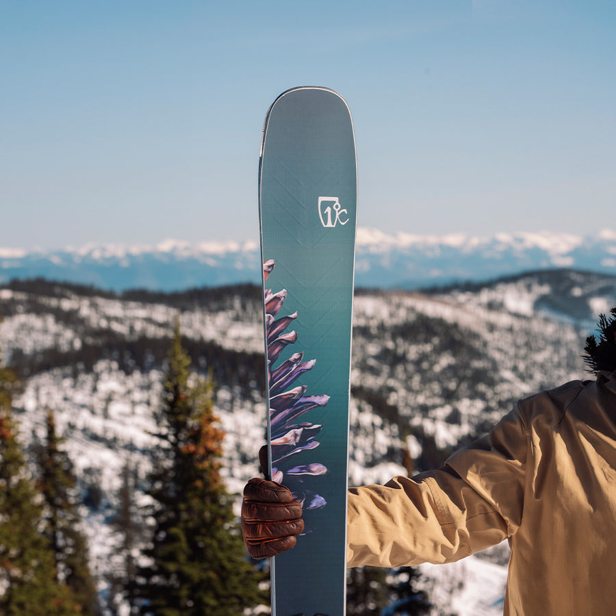 Dressed in a tan jacket, a skier proudly holds up an Icelantic Skis Torrent 102—featuring its distinctive handprint design—while standing atop a snowy mountain framed by pine trees and sweeping mountain ranges, embodying the adventurous spirit of an expert ready to carve fresh tracks.
