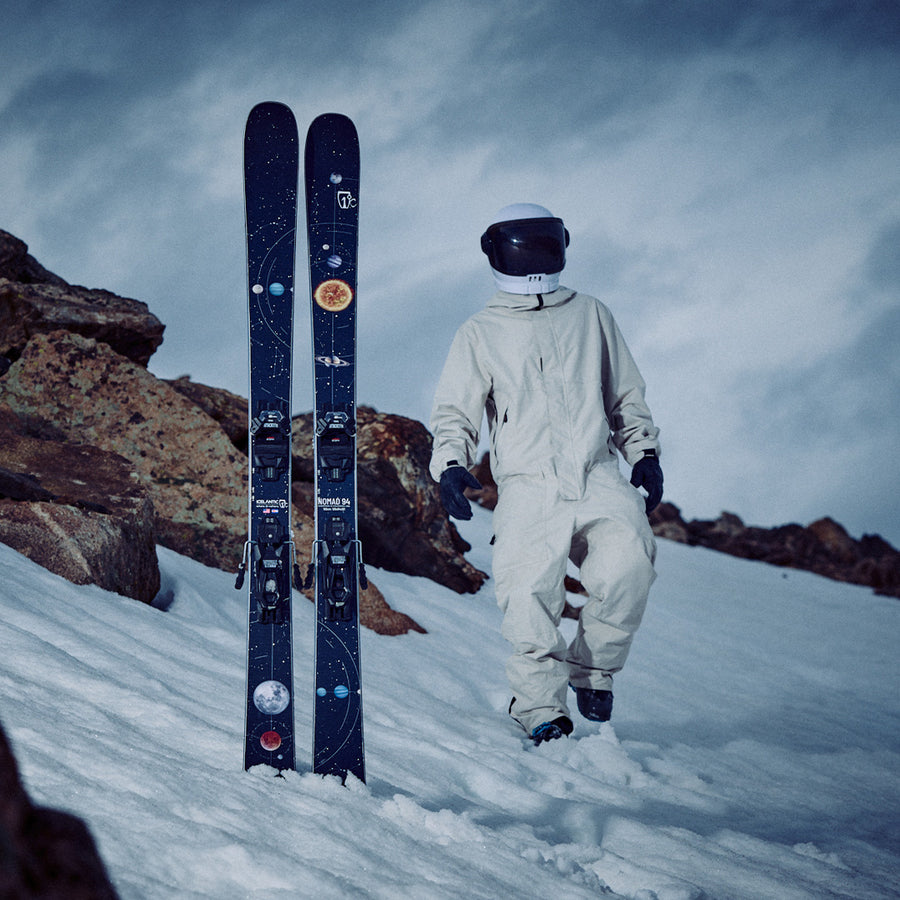 Clad in a white spacesuit, a person stands on snowy terrain near rocks beside a pair of Icelantic Skis Nomad 94 all-mountain skis adorned with planets and stars, under a cloudy sky.