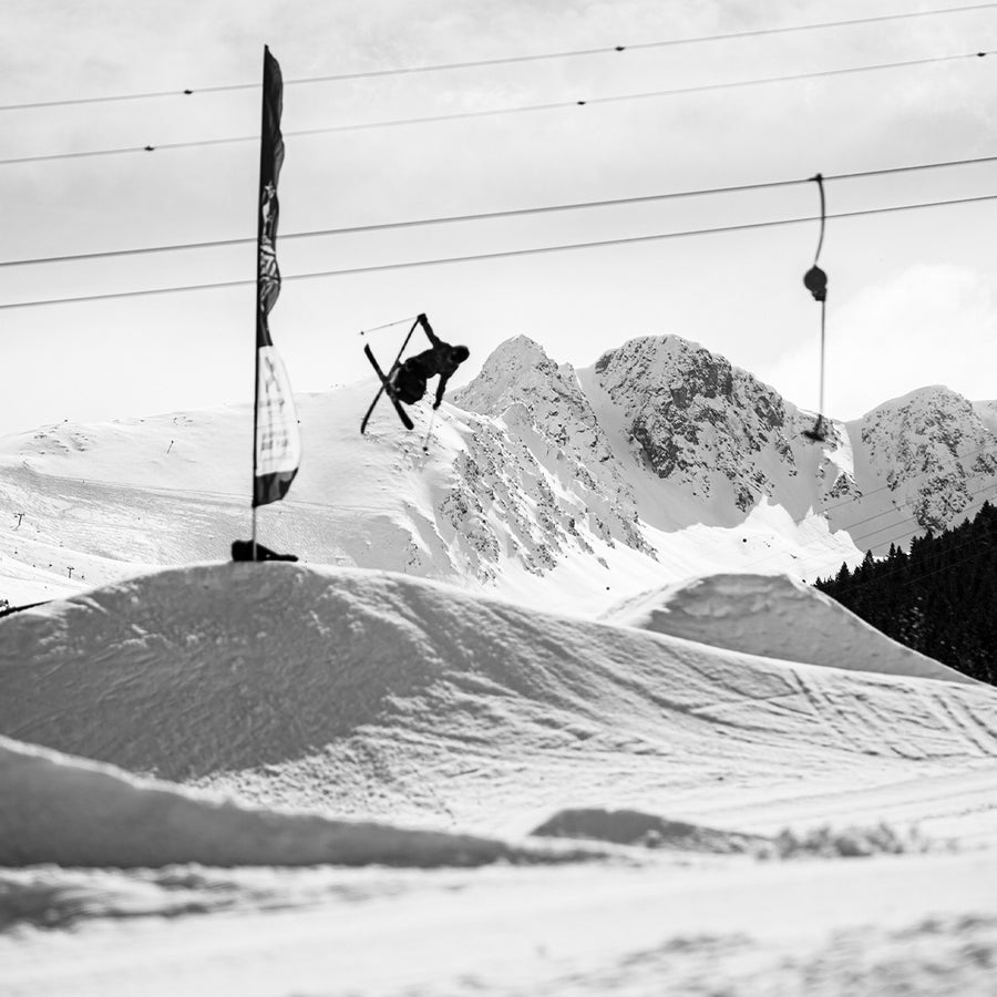 A skier launches off a snowy jump in a mountainous terrain park on Icelantic Skis Nomad 94 Whiteout freestyle skis, with ski lifts and snow-covered peaks in the background.