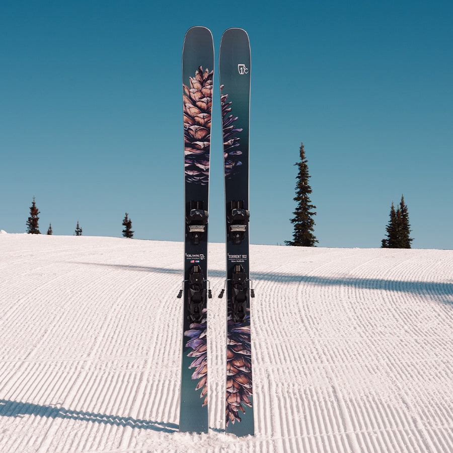 A pair of Icelantic Skis Torrent 102, adorned with bold pinecone graphics and engineered with the advanced V-12 Carbon Matrix, stand upright on a groomed snowy slope beneath a clear blue sky, while evergreen trees frame the scene in the background. Ideal for advanced skiers who demand top-tier performance and standout style.