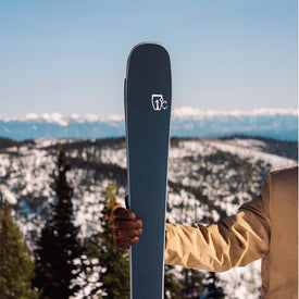 Someone wearing a tan jacket and brown glove holds the Icelantic Skis Torrent 88 ski upright, with snowy mountains and pine trees in the background under a clear blue sky—ready for agile rides and all-mountain performance.