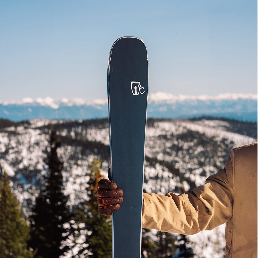 Someone wearing a tan jacket and brown glove holds the Icelantic Skis Torrent 88 ski upright, with snowy mountains and pine trees in the background under a clear blue sky—ready for agile rides and all-mountain performance.
