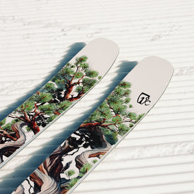 A pair of white Icelantic Skis Torrent 96 all-mountain skis, adorned with intricate tree and pine branch artwork, rest on groomed, ridged snow under bright sunlight.