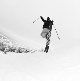 A skier in mid-air performs a trick on a snowy slope, holding ski poles outward, highlighting the agility of the Icelantic Skis Nomad 100 Whiteout all-terrain skis. Snow-covered trees and a mountain landscape serve as the backdrop in this black-and-white image.