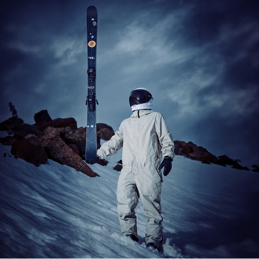 An astronaut in a white suit stands on a snowy slope at dusk, holding a pair of Icelantic Skis’ Nomad 94 all-mountain skis upright. Rocky terrain stretches in the background under a dark, cloudy sky.