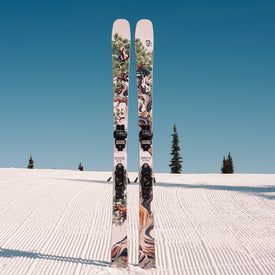 Two Icelantic Skis Torrent 96 all-mountain skis featuring artistic tree graphics stand upright on a freshly groomed, snow-covered slope beneath a clear blue sky, with several pine trees visible in the background.