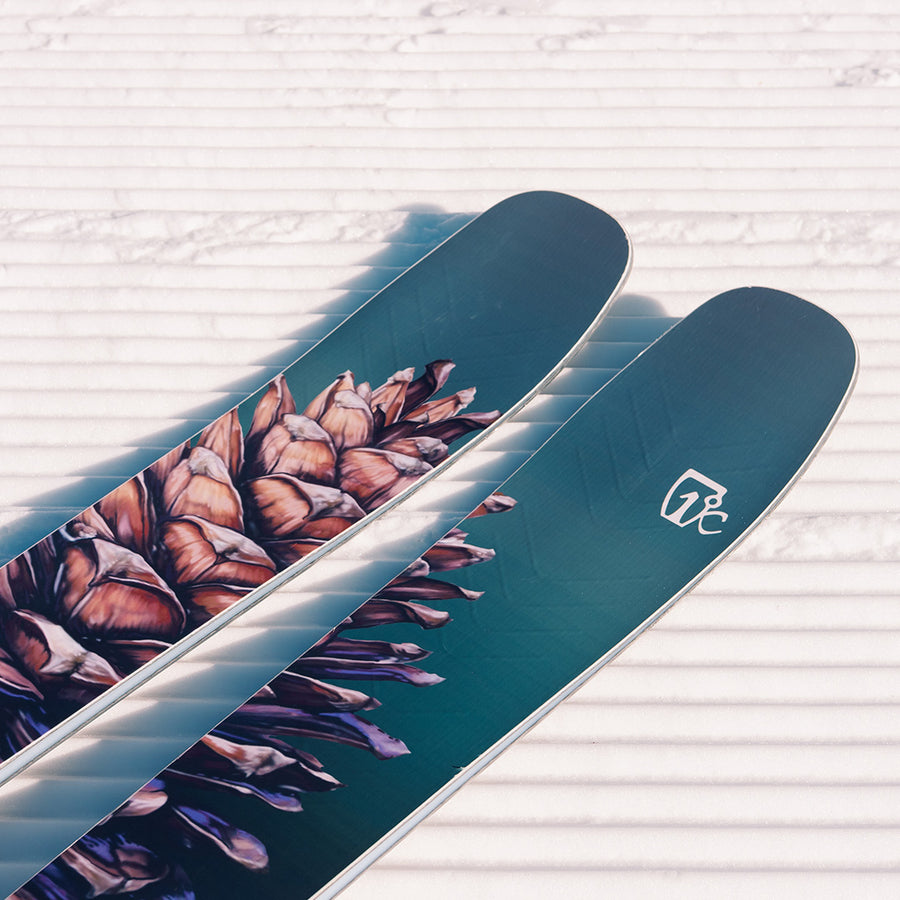 A close-up of two Icelantic Skis Torrent 102s with a distinctive pinecone graphic, featuring the advanced V-12 Carbon Matrix design, resting on freshly groomed snow with crisp parallel lines—ideal for the advanced skier in search of peak performance.