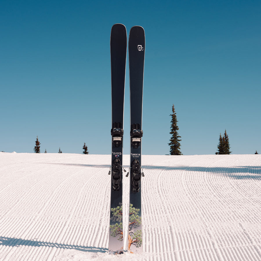 A pair of Icelantic Skis Torrent 88, acclaimed for their all mountain performance, standing upright on a freshly groomed snowy slope with a clear blue sky and a few evergreen trees in the background.