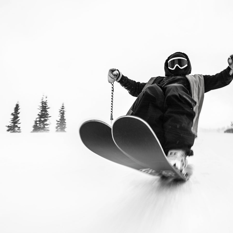 Clad in winter gear and goggles, a skier speeds downhill on Icelantic Skis’ Nomad 100 Whiteout, captured from a low angle with motion blur; snow-covered ground and pine trees fill the background, highlighting the all-terrain performance and agility of these skis.