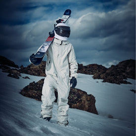 Clad in a white snowsuit and reflective helmet, an individual stands on a snowy slope with their Icelantic Skis Nomad 100 all terrain ski slung over their shoulder. Rocky terrain and dramatic clouds create a striking backdrop.