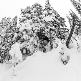 A skier launches through snow-covered trees on a powdery slope, riding the versatile Icelantic Skis Nomad 106 Whiteout—an ideal freeride ski for intermediate to expert skiers. The black-and-white scene captures the exhilarating thrill of the moment.