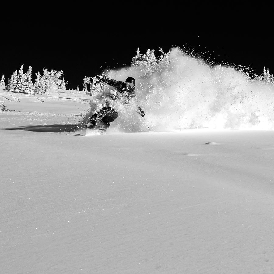 A snowboarder carves through deep powder snow, sending up a dramatic spray amidst a snow-laden landscape dotted with trees beneath a clear sky. Captured in striking black and white, this dynamic scene embodies the exhilaration that intermediate to expert skiers can experience while tackling challenging freeride terrain on the Icelantic Skis Nomad 106 Whiteout.