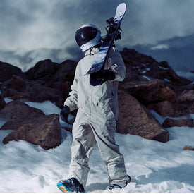 A person in a white spacesuit and helmet stands on snowy ground, holding an Icelantic Skis Nomad 106 freeride ski, with rocky mountains in the background.