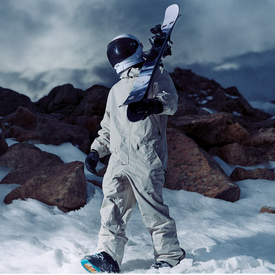 A person in a white spacesuit and helmet stands on snowy ground, holding an Icelantic Skis Nomad 106 freeride ski, with rocky mountains in the background.