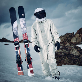 A person in a white spacesuit stands on snow beside upright Icelantic Skis Nomad 100 all-mountain skis, which showcase an eye-catching moon graphic. Rocky terrain and a cloudy sky form the backdrop, evoking the adventurous spirit of freestyle skiing that pushes the limits of earthly exploration.