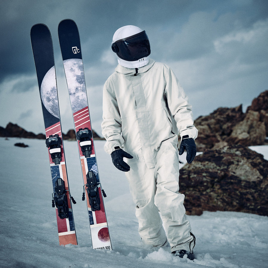 A person in a white spacesuit stands on snow beside upright Icelantic Skis Nomad 100 all-mountain skis, which showcase an eye-catching moon graphic. Rocky terrain and a cloudy sky form the backdrop, evoking the adventurous spirit of freestyle skiing that pushes the limits of earthly exploration.