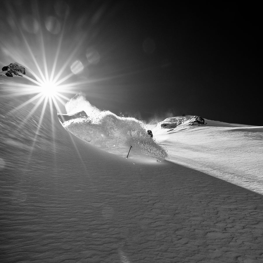 A skier carves through deep snow on a mountain slope under a radiant sunburst, sending up a trail of powder and dramatic shadows in this striking black and white photo—an exhilarating moment captured on the Icelantic Skis Nomad 112 Whiteout, perfect for advanced skiers seeking the thrill of freeride adventures.