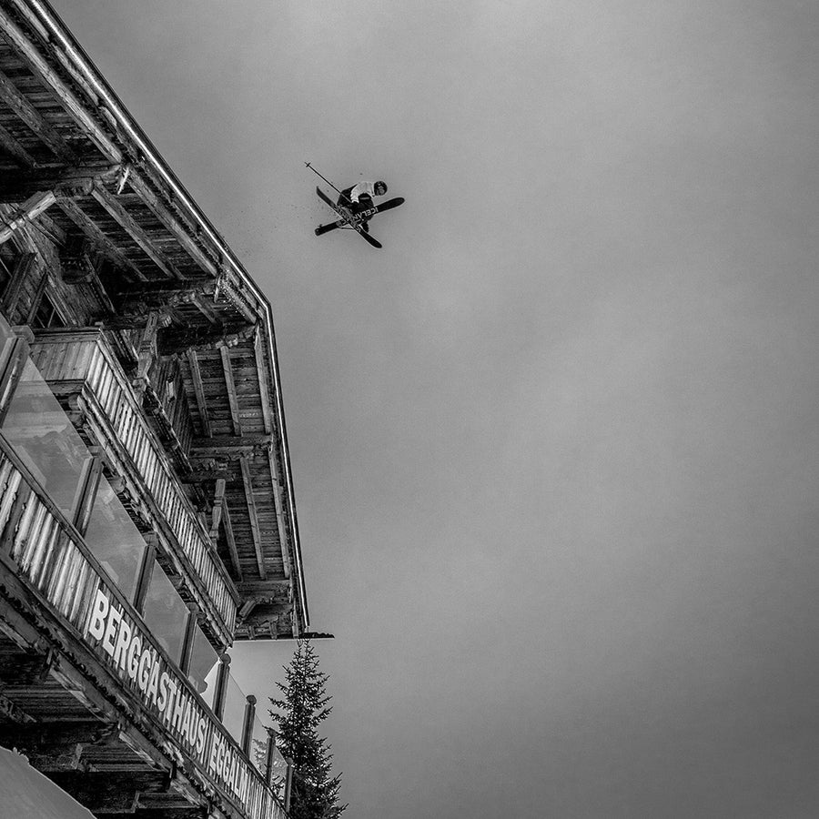 A skier launches high beside a wooden alpine lodge, captured mid-air against a cloudy sky while demonstrating freeride technique on the Icelantic Skis Nomad 112 Whiteout. The black and white scene highlights both the dynamic jump and the sleek design of the skis.