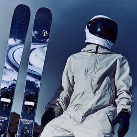 Wearing a spacesuit, a person stands beside two Icelantic Skis Nomad 106 freeride skis, which feature space-themed designs with planets and stars, set against a dark sky background.