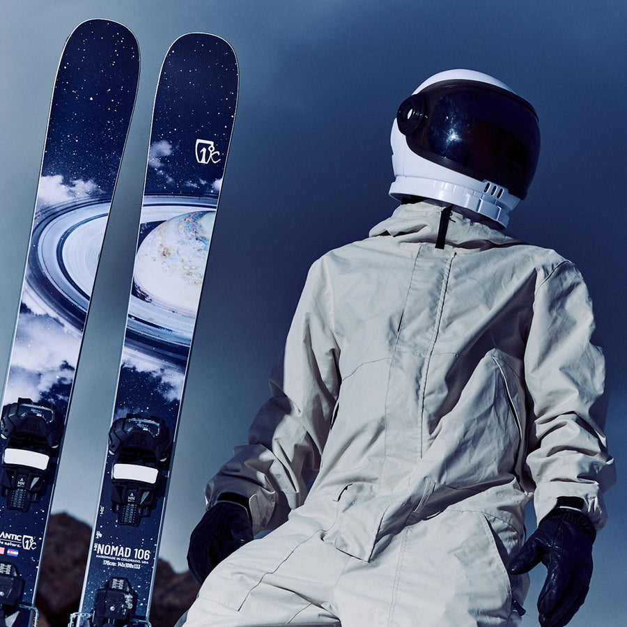 Wearing a spacesuit, a person stands beside two Icelantic Skis Nomad 106 freeride skis, which feature space-themed designs with planets and stars, set against a dark sky background.